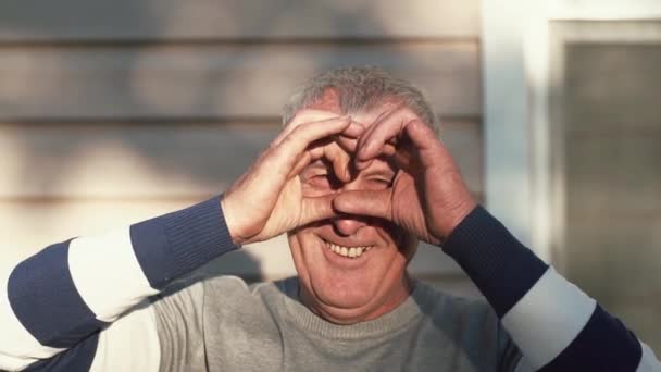 An old man looks into camera, makes a shape of glasses with his fingers, smiles joyfully. Slow mo — Stock Video
