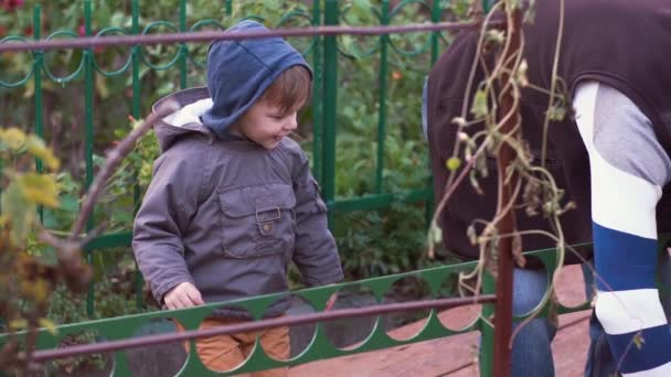 Little boy looks at an old man gardening. The boy touches the fence, makes silly face. Slow mo — Stock Video