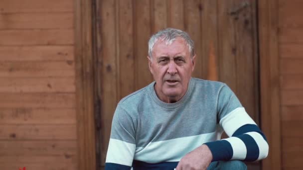 An ageing man sits near a wooden house and looks into camera. Slow mo — Stock Video