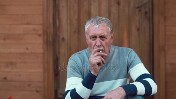 Un vecchio si siede vicino a una casa di legno, guarda nella macchina fotografica e fuma. Lento mo — Video Stock