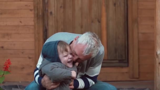 Старий сидить біля будинку, грає зі своїми онуками, цілує і обіймає їх. Діти сміються і розважаються. Повільний mo — стокове відео