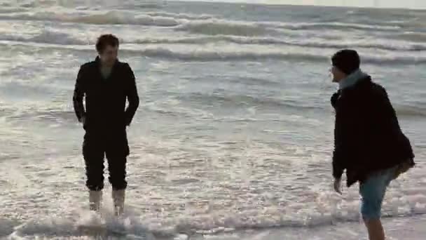 Two young guys are soaking their feet in the waves of the sea smiling and talking to each other. — Stock Video