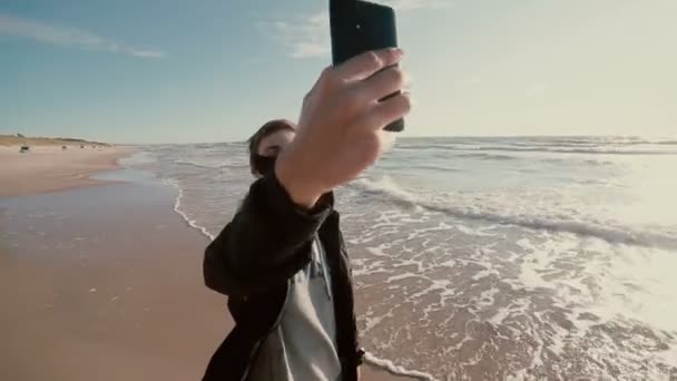 Een jonge gelukkige kerel is permanent blote voeten in het water met zijn opgerolde spijkerbroek en proberen te maken een selfie. — Stockvideo