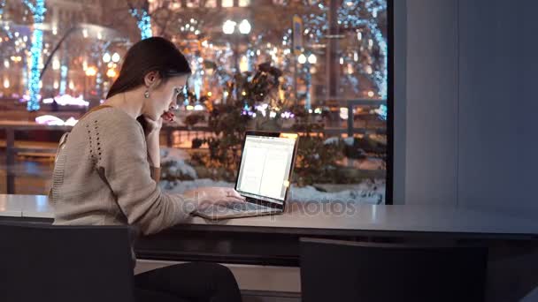 Atractiva joven mujer de negocios que trabaja en el portátil sentado en el bar, fuera de la ciudad noche de invierno decorado para Navidad — Vídeos de Stock