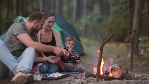 Descanso familiar en la naturaleza. Esposo y esposa con su pequeño hijo están cocinando malvaviscos en fuego abierto y comiéndolos. . — Vídeos de Stock