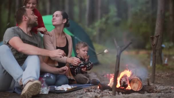 Perhe lepää luonnossa pienen poikansa ja tyttärensä kanssa, he keittävät vaahtokarkkeja avotulella ja syövät ne. . — kuvapankkivideo