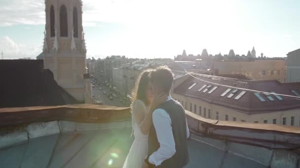 Gelukkige jonge mooie stijlvolle paar bruid en bruidegom knuffelen zachtjes op het dak bij zonsondergang city straat op de achtergrond — Stockvideo