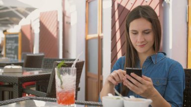 Esmer kız akıllı telefon süre dışında oturan bir kafede, içme ve saman tüp soğuk bir içecek keyfini kullanır. 4k,
