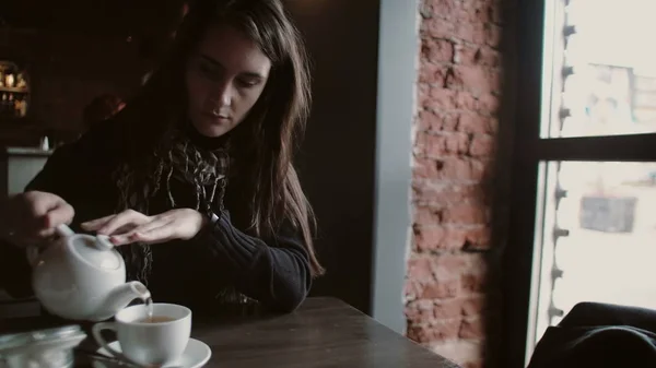 Молодая девушка наливает чай из чайника в кружку, сидя в современном кафе — стоковое фото