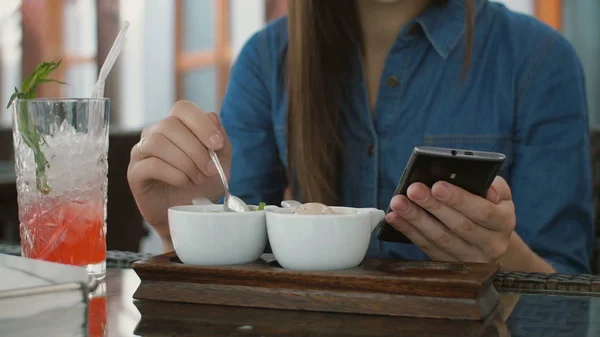 Brunette meisje gebruikt slimme telefoon zittend buiten in een café, het eten van ijs. 4k, — Stockfoto