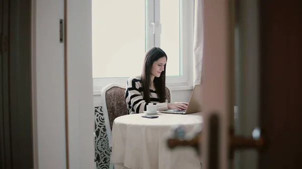 Kobieta używa laptopa i korzystania z kawy rano w jasnej jadalni. Zobacz przez otwarte drzwi — Zdjęcie stockowe