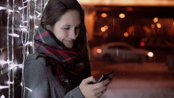Jovem mulher atraente usando smartphone na neve caindo na noite de Natal de pé perto de luzes parede , — Fotografia de Stock
