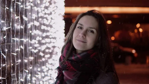 Молодая привлекательная женщина в падающем снегу в Рождественскую ночь, глядя на огни камеры на заднем плане — стоковое фото