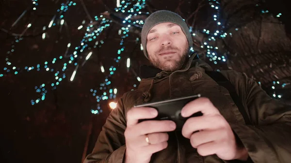 Όμορφος νεαρός χρησιμοποιώντας το smartphone στο Χριστούγεννα νύχτα στέκεται κάτω από ένα δέντρο διακοσμημένο με αφρώδη φώτα — Φωτογραφία Αρχείου