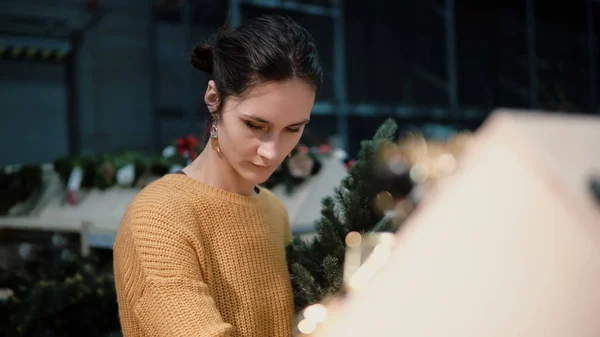 Giovane ragazza bruna attraente al negozio sceglie un albero di Natale artificiale e giocattoli, decorazione di Natale . — Foto Stock
