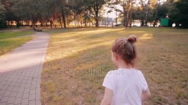 Neşeli küçük bir kız bir parkta bir yolda bir güneşli yaz günü çalışır. Arkadan Görünüm, yavaş mo