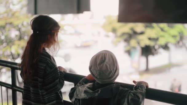 Un niño y una niña apoyados en una barandilla de puente, mirando hacia la calle de la ciudad. Vista trasera — Vídeo de stock