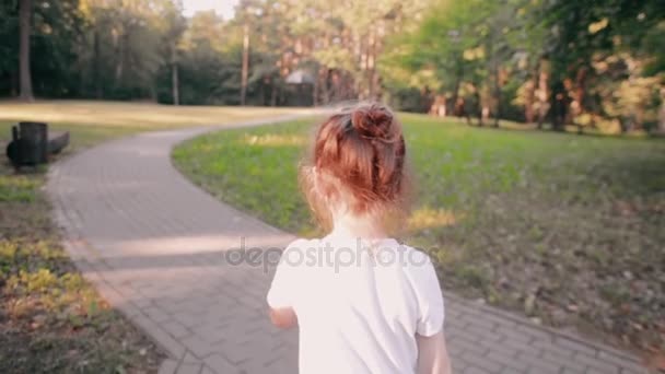 Liten flicka som går på en väg i en park. En bulle av ljust hår har guld glöd i solen. Långsamma mo, bakifrån — Stockvideo