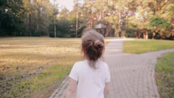 Маленькая девочка идет по дороге в парке. У пучка светлых волос золотое сияние на солнце. Медленно, вид сзади — стоковое видео