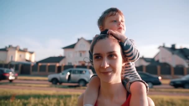 A mãe e o filho passam algum tempo juntos. Menino bonito senta-se em seus ombros mães, ela sorri feliz. O sol brilha. Devagar. — Vídeo de Stock