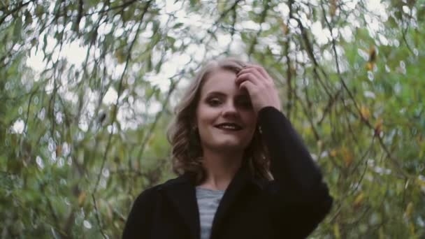 Стильная красивая молодая блондинка позирует перед камерой на фоне листьев деревьев, замедленное движение — стоковое видео
