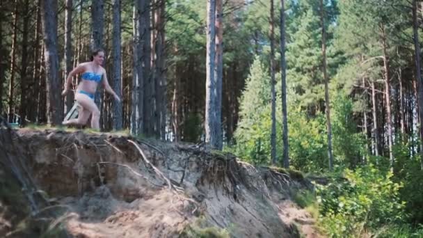 Lively woman in a bathing suit enjoys time in the nature. She runs and jumps into water. Forest at the background. — Stock Video