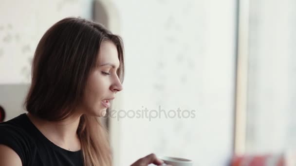 Närbild på en brunett kvinnans ansikte med en kopp kaffe eller te. Hon talar, leenden, har god tid. — Stockvideo