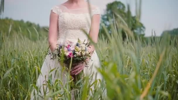 A beautiful young bride smelling her wedding bouquet standing in a wheat field. Pretty lacy wedding dress on her. — Stock Video