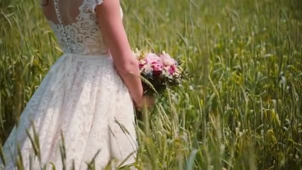 Close-up de um belo vestido de noiva rendada em uma noiva de pé em um campo de trigo com um buquê em suas mãos . — Vídeo de Stock