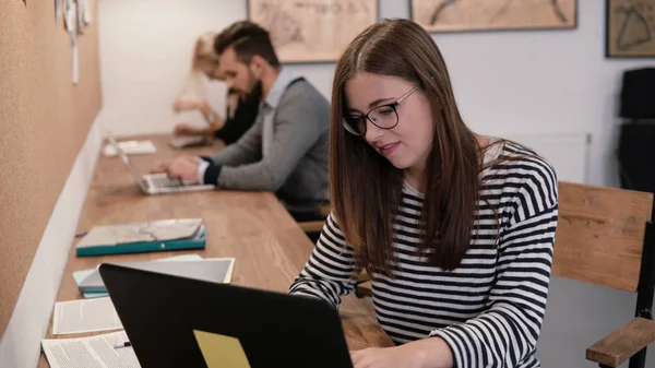 Joven chica atractiva está utilizando un ordenador portátil, trabajando con documentos, tomando notas en la moderna oficina de inicio — Foto de Stock