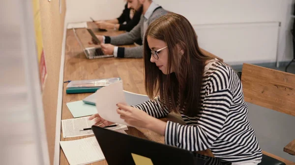 Joven chica atractiva está utilizando un ordenador portátil, trabajando con documentos, tomando notas en la moderna oficina de inicio — Foto de Stock