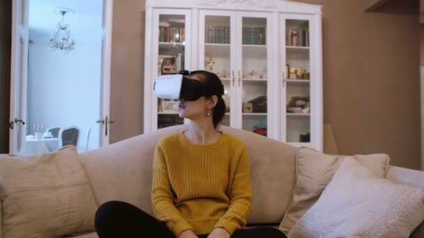 Mulher bonita sentada no sofá na sala de estar e usando fone de ouvido VR. 4K — Vídeo de Stock