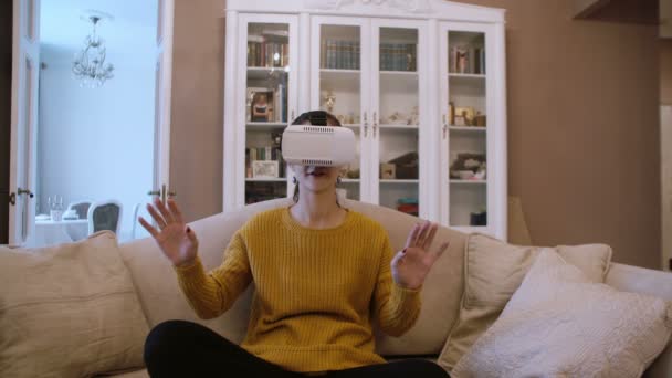 Hermosa joven sentada en el sofá en la sala de estar y usando auriculares VR. 4K — Vídeo de stock