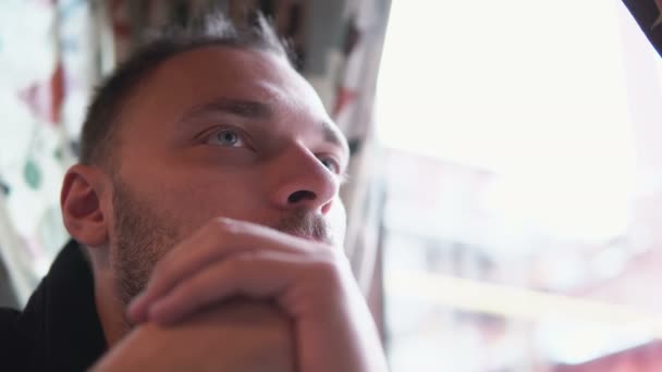 Joven hombre guapo sentado en la cafetería cerca de la ventana y mirar a la distancia. Tomando de la mano delante de la cara. Primer plano, 4K — Vídeos de Stock