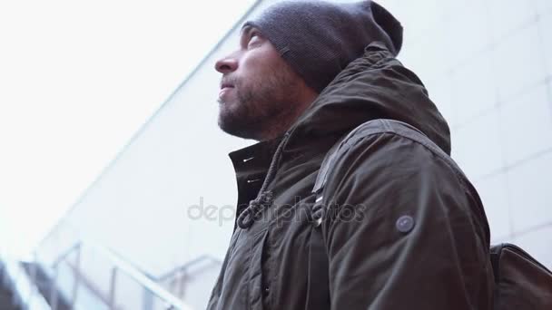Молодой привлекательный мужчина в куртке и шляпе стоит один на улице и смотрит вокруг в холодный день. 4K — стоковое видео