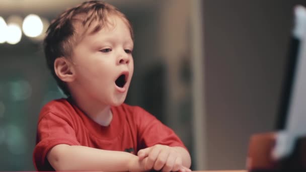 Niño pequeño con camisa roja sentado delante de la tableta y bostezos — Vídeos de Stock