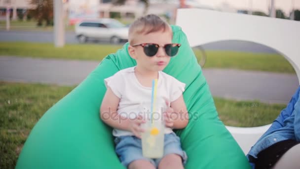 Grappig jongetje in een grote zonnebril zittend in bean bag stoel in het park in zomerdag. Houd een glas met rietjes. — Stockvideo