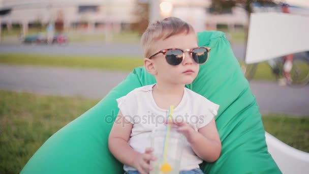 Carino bambino in un grande occhiali da sole divertenti seduti in poltrona sacchetto di fagioli al parco in giorno d'estate. Tenendo un bicchiere con cannucce — Video Stock