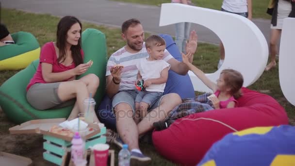 Glad ung familj med två barn vilar på en picknick i parken. Pojke och flicka äter pizza, sitter i bean bag stolar — Stockvideo