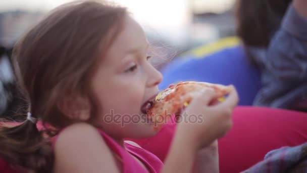 Hermosa niña linda sentada en sillas bolsa de frijoles y comer pizza en el parque en el día de verano. Vista lateral . — Vídeos de Stock