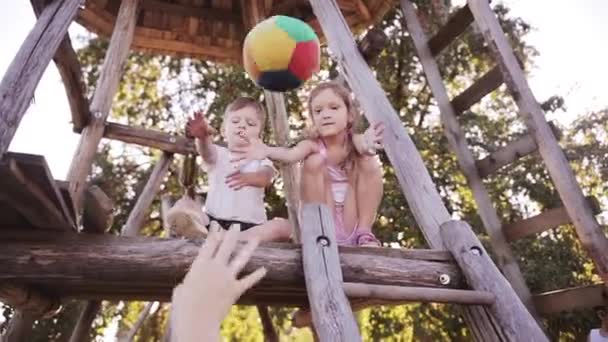 Gyönyörű mosolygó lány és egy fiú testvére játszani labdát, kültéri fa-ház, valamint a szülő a napsütéses napon — Stock videók