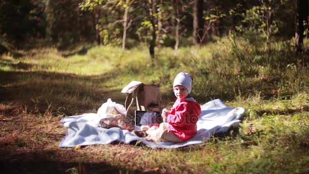Niño lindo tienen picnic en el bosque en otoño, sentado en la hierba, mira a la cámara. Vista lateral . — Vídeos de Stock