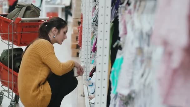 Jeune belle femme accroupie au supermarché et choisissant des vêtements de bébé à la vitrine. Près du caddie féminin. 4K — Video