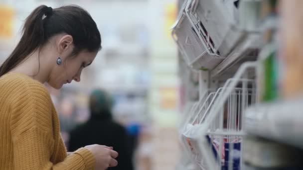 Молодая красивая женщина в супермаркете стоит перед полками и смотрит на что-то, затем поворачивается и уходит. 4K — стоковое видео