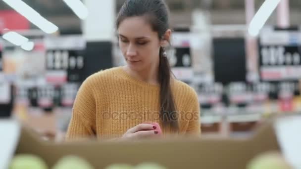 Vista frontal da jovem mulher escolhendo uma maçã verde no supermercado. A menina jovem considera frutos. 4K — Vídeo de Stock
