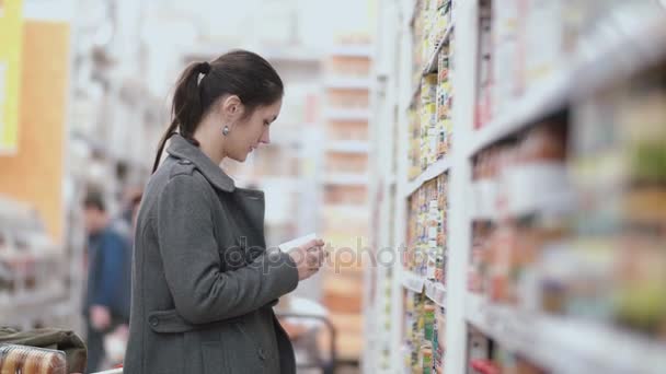 Joven hermosa mujer de pie delante de los estantes con la tienda de comestibles, sosteniendo uno puede y tratar de elegir en el supermercado. 4K — Vídeo de stock