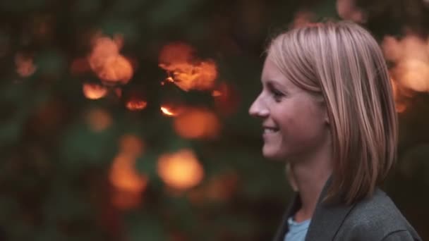 Giovane bella donna in piedi al parco nei raggi del sole al tramonto, sorridente, godendo della natura. Primo piano. Vista laterale . — Video Stock