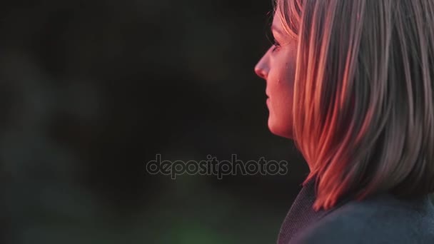 Ελκυστική νεαρή γυναίκα στέκεται στο πάρκο στις ακτίνες του ήλιου ρύθμιση και ψάχνει σε απόσταση. Γκρο πλαν θέα — Αρχείο Βίντεο