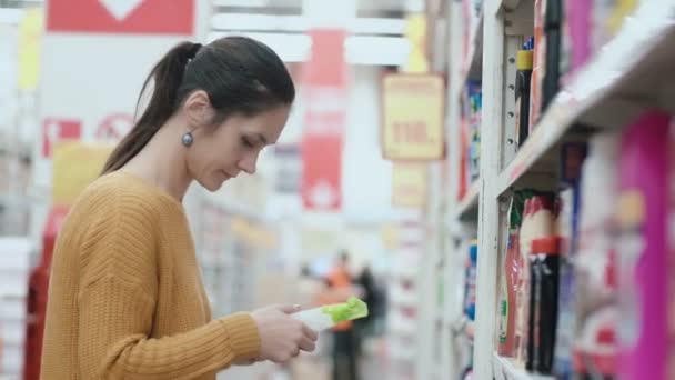 Young beautiful woman standing in front of shelves with household chemicals, holding one and try to choose. 4K — Stock Video