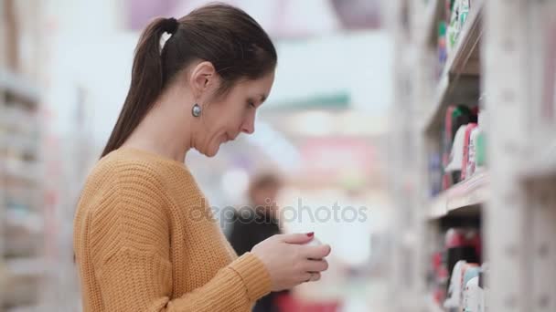Молодая привлекательная женщина стоит перед полками с дезодорантом, держа два и попытаться выбрать. 4K — стоковое видео